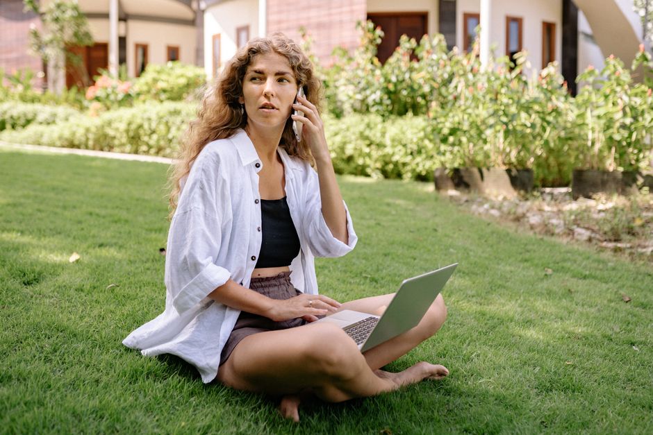Uma mulher vestindo roupas casuais trabalha remotamente em um laptop e celular enquanto está sentada na grama.