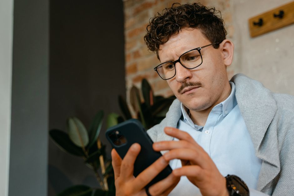 Um homem concentrado com óculos usando um smartphone em ambientes internos ao lado de uma parede de tijolos.