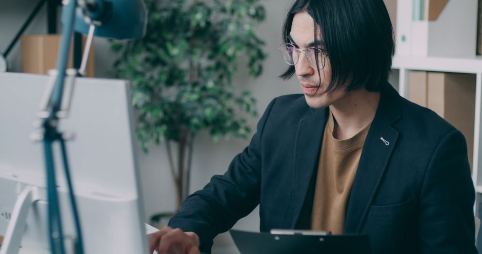 Homem de óculos concentrado no trabalho em uma mesa de computador em um ambiente de escritório contemporâneo.