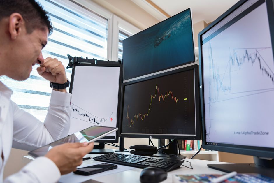Trader com camisa branca analisando gráficos de ações em vários monitores durante o dia em um ambiente de escritório.